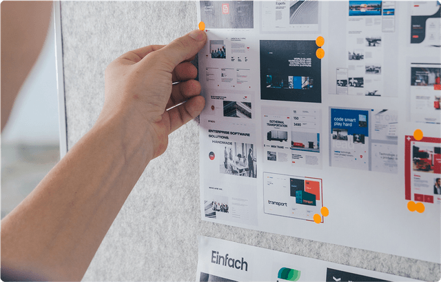 A person pins a sheet of web design layouts on a board, surrounded by orange pushpins and other design sheets.