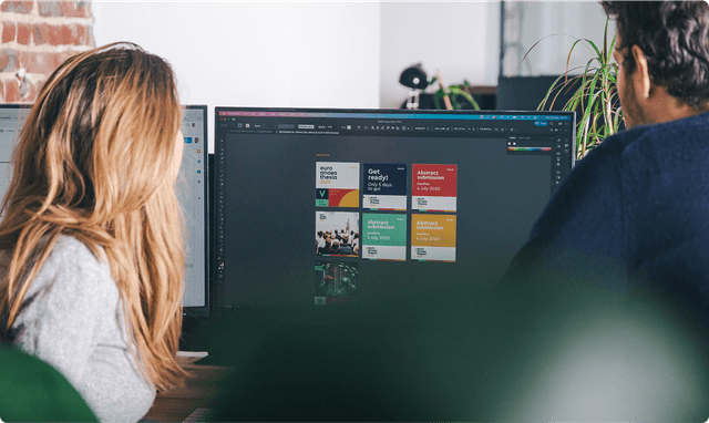 Two people working at a desk, viewing design software on a large monitor displaying colorful graphics and text.