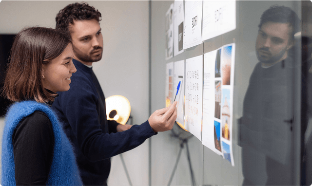 Two people discussing and pointing at documents on a glass wall, with a lamp in the background.