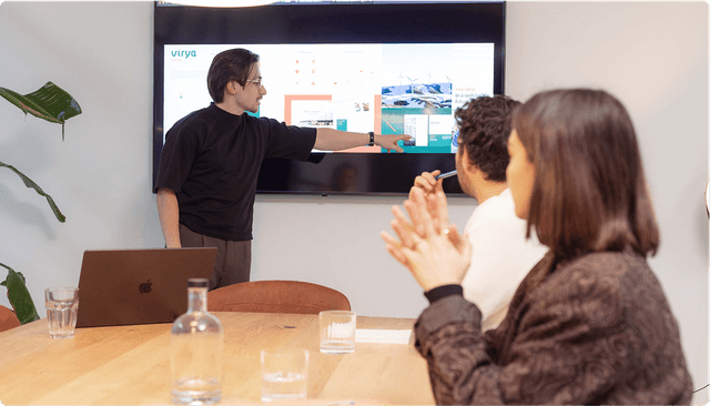A person presents slides on a screen to two seated individuals in a meeting room with a wooden table and a plant in the corner.