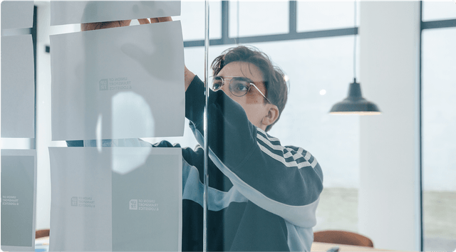 Person in glasses and a striped sweater attaching papers to a glass wall in a bright room with modern decor.