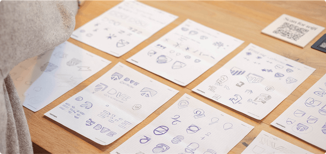 Sheets of paper on a wooden table displaying various hand-drawn logo sketches and designs in blue ink.