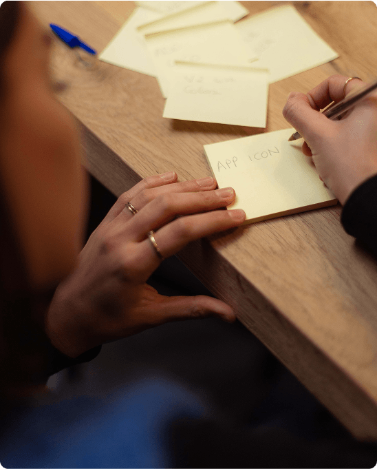 Person writing "APP ICON" on a sticky note at a wooden desk, surrounded by other notes and a pen.
