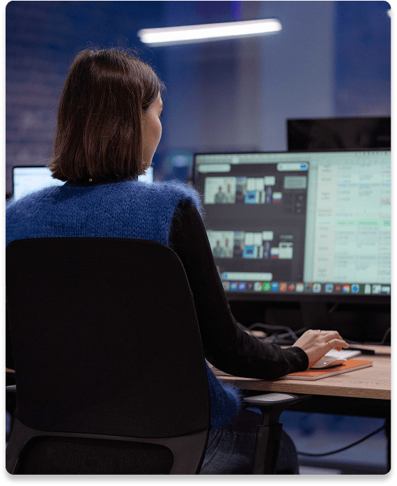 Person with short hair working on a computer with multiple screens displaying various data and images in a dimly lit office.