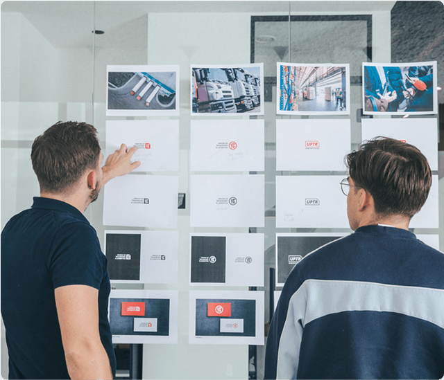 Two people examining a wall display of printed images and designs, discussing various layouts in a modern office setting.