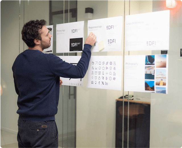 Man reviewing design documents on a glass wall, featuring logos, icons, and photography samples.