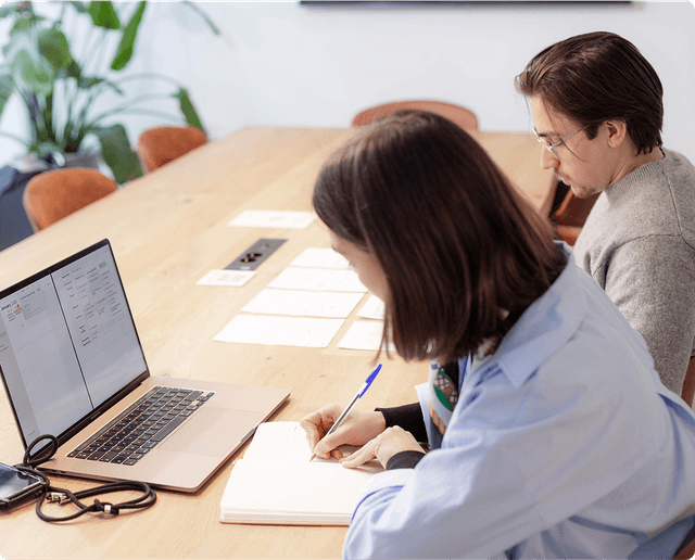 Two people working at a wooden table with a laptop, papers, and notebooks. One is writing, the other is focused on the screen.