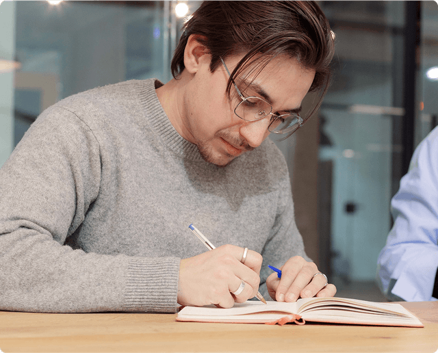 A person with glasses and a gray sweater writes in a notebook at a wooden table in a modern office setting.