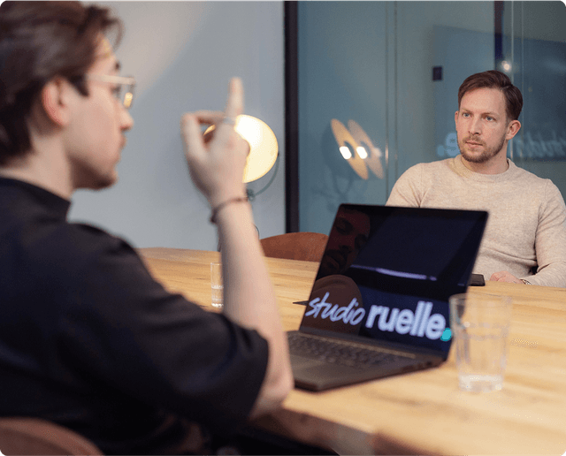 Two people in a meeting, one gesturing while the other listens. A laptop with "studio ruelle" on the screen is on the table.