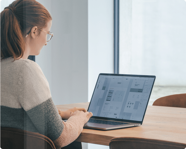 Architecture Person with glasses and red hair working on a laptop at a wooden table, viewing a design software interface. Large window in the background.