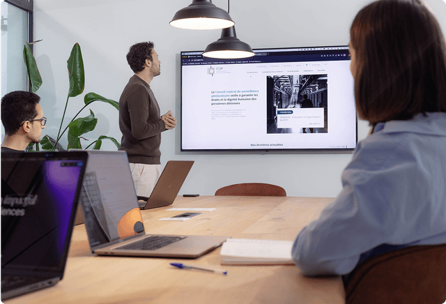 Prototype Three people in a meeting room, one presenting on a large screen displaying a website. Laptops are open on the table.